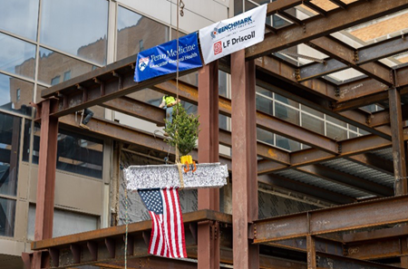 A steel beam is lifted into place in the Emergency Department project and attached to the beam is the American flag and a Christmas tree